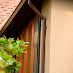 A dark brown seamless gutter downspout contrasting the beige exterior of the home 